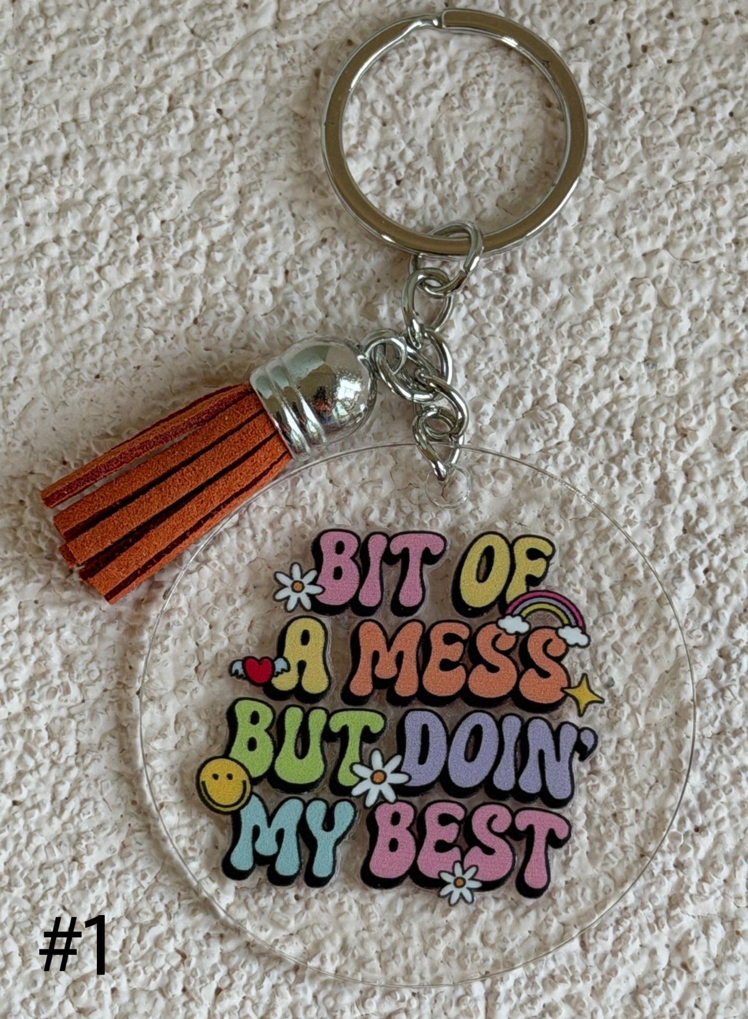 A round clear acrylic keychain featuring colorful retro-style bubble lettering that reads 'BIT OF A MESS BUT DOIN' MY BEST' with small doodle accents. The keychain includes a silver metal key ring, chain, and a brown leather tassel.