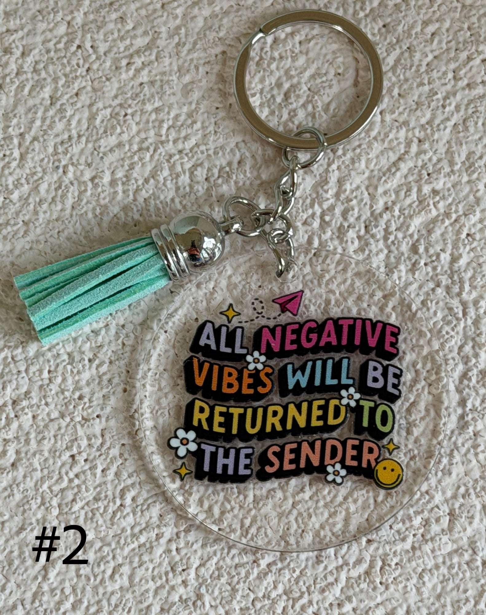A clear acrylic keychain with a mint green faux-leather tassel and a silver metal key ring. The acrylic charm features the text 'ALL NEGATIVE VIBES WILL BE RETURNED TO THE SENDER' in colorful bubble letters with small decorative doodles.
