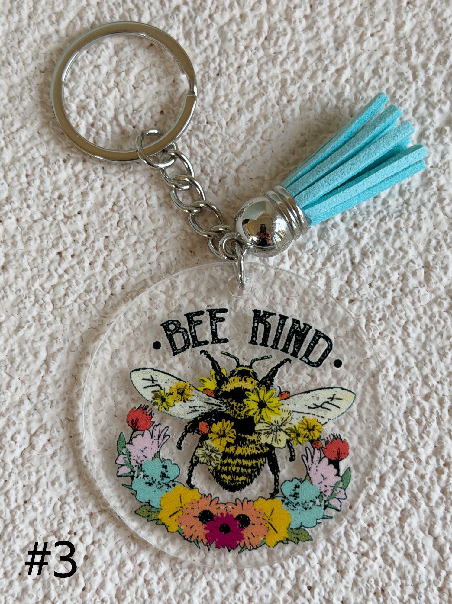 A round acrylic keychain featuring a colorful bee and flower illustration with the text 'BEE KIND.' and a light blue tassel, shown on a textured white background.