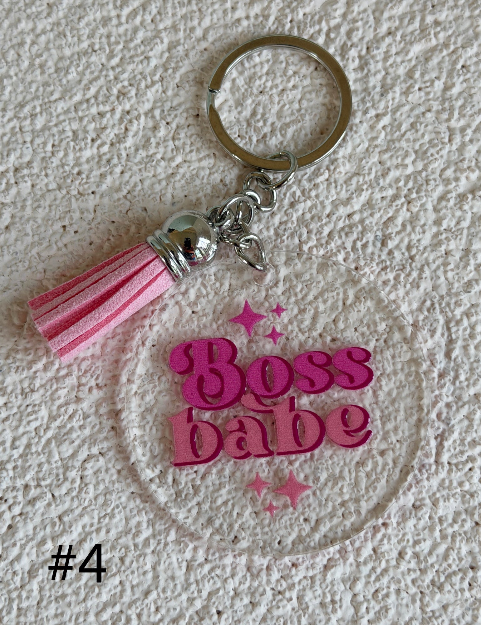 A pink and silver keychain featuring a clear acrylic disc with the words 'Boss babe' in pink glittery letters and small pink stars, alongside a pink and white striped tassel, shown on a textured white background.