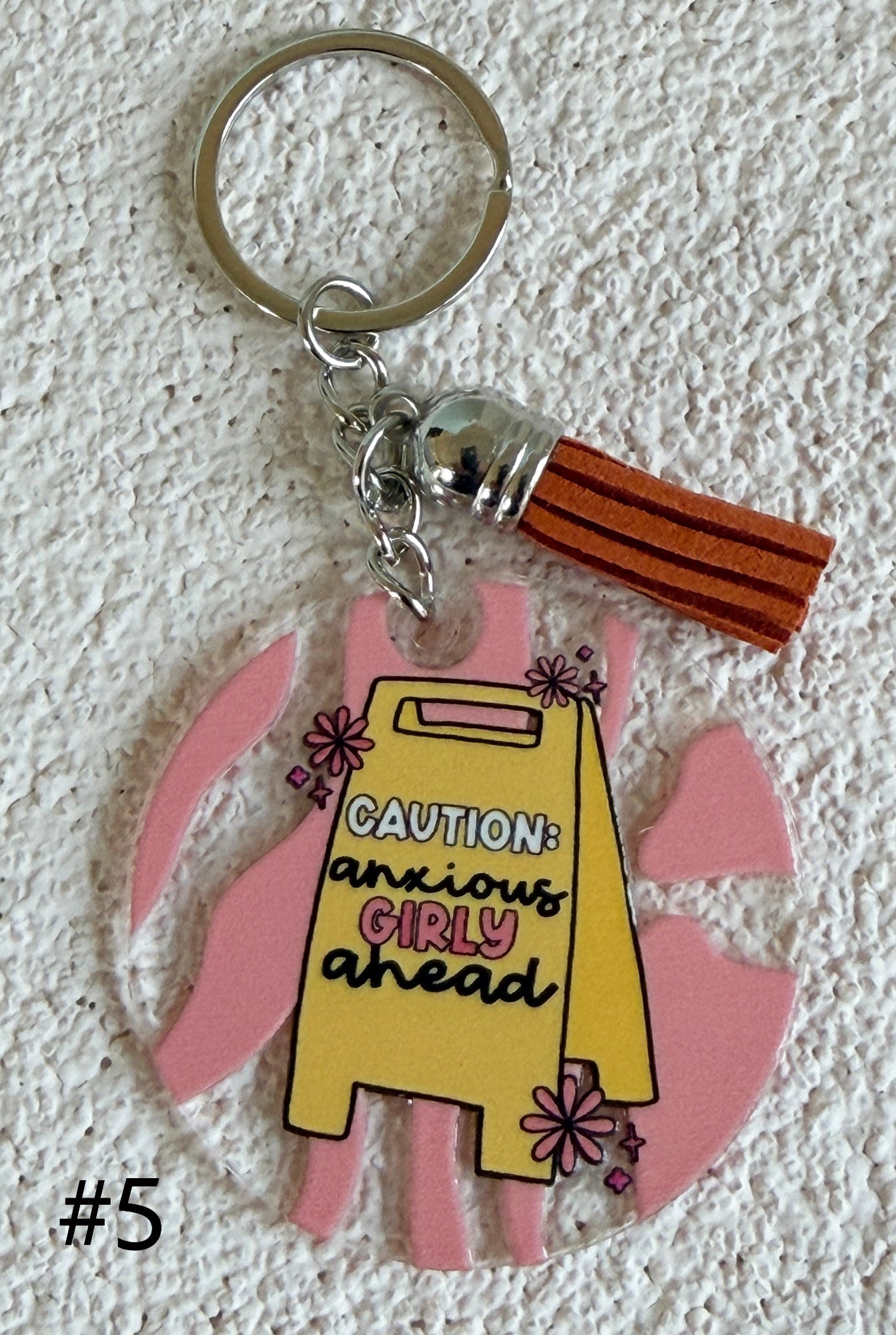 A novelty keychain featuring a yellow apron charm with the text 'CAUTION: anxious GIRLY ahead' in pink and black lettering, surrounded by pink bunny ears and a brown leather tassel, displayed on a textured white background.