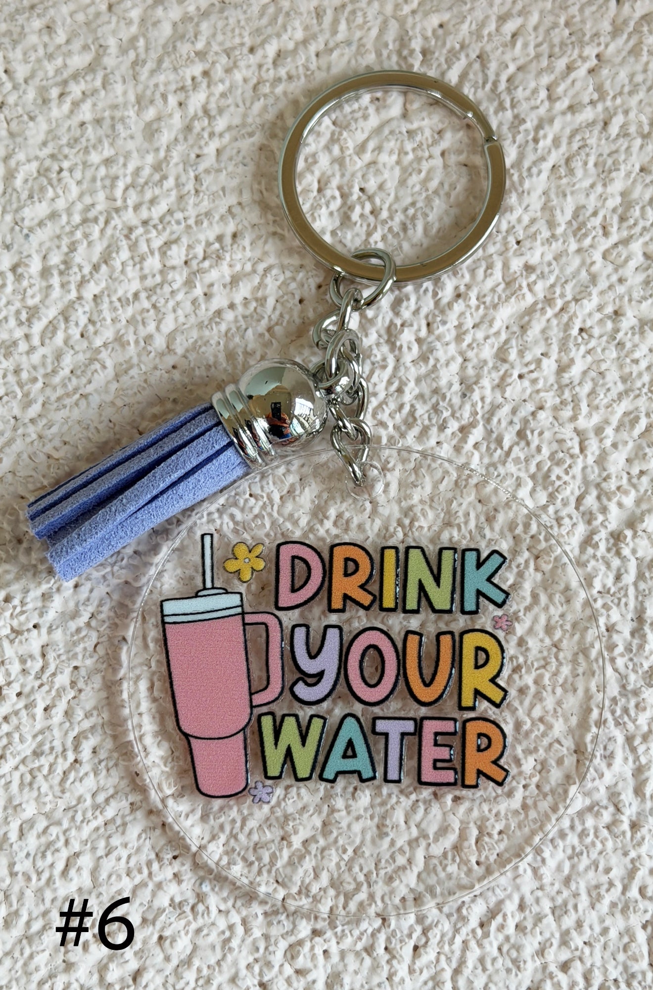 A clear acrylic keychain featuring a tassel charm and a circular charm with the text 'DRINK YOUR WATER' in pastel colors next to a pink iced drink illustration. The keychain is photographed on a textured off-white surface.