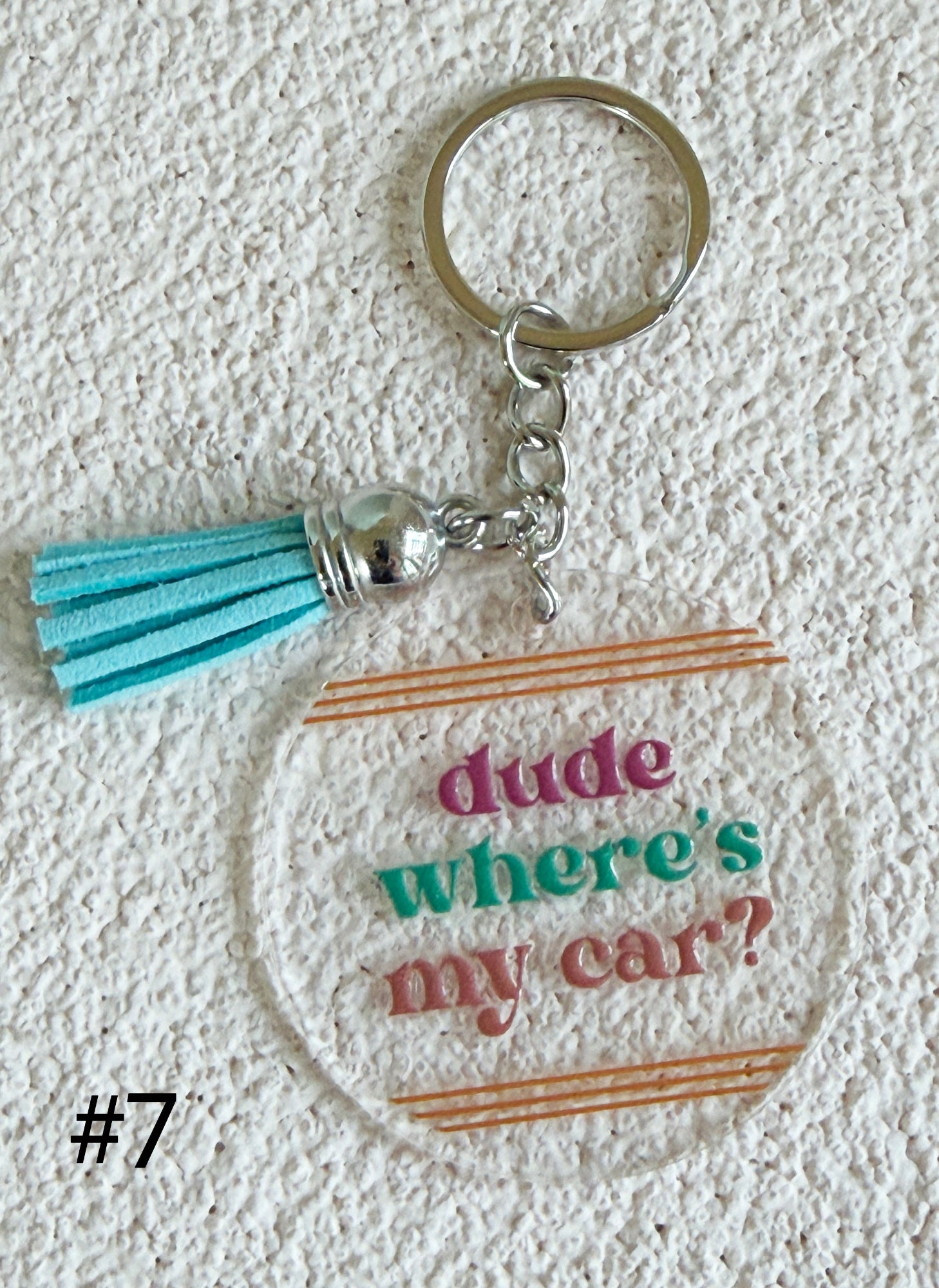A clear acrylic keychain with a silver metal split ring and tassel charm, featuring the text 'dude where's my car?' in pink, green, and brown lettering on a textured white background.