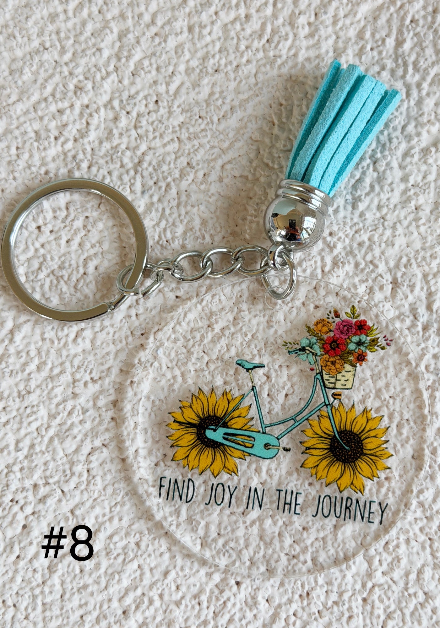 A round acrylic keychain featuring a turquoise bicycle with yellow sunflowers and a basket of flowers, with the text 'FIND JOY IN THE JOURNEY' and a turquoise tassel on a textured white background.