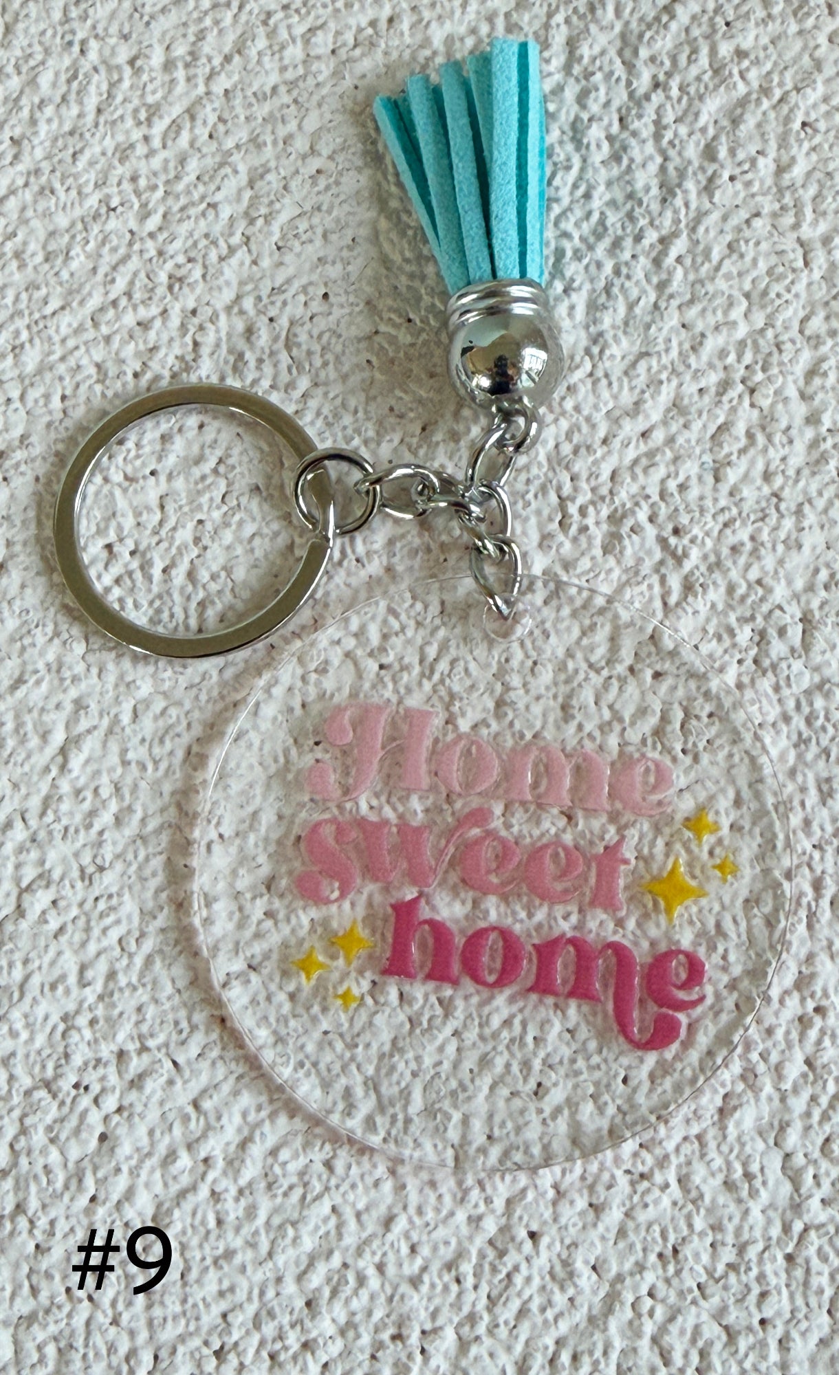 A clear acrylic keychain featuring the phrase 'Home sweet home' in pink script with yellow star accents, attached to a silver split ring and topped with a turquoise tassel, displayed on a textured white background.