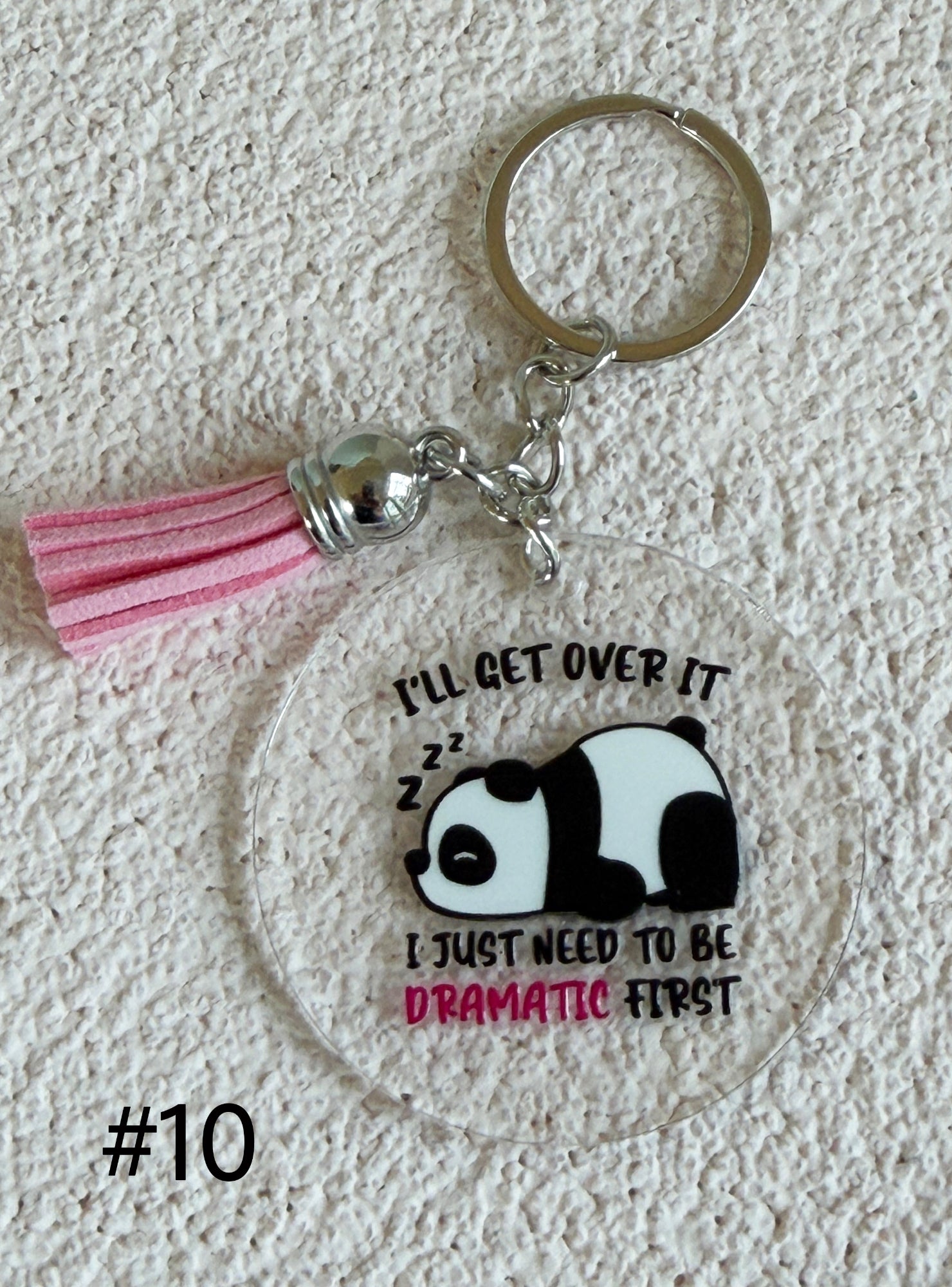A round acrylic keychain featuring a sleeping panda graphic with the text 'I'LL GET OVER IT I JUST NEED TO BE DRAMATIC FIRST' in black and pink lettering. The keychain includes a pink tassel, silver metal chain, and a silver split key ring, displayed on a textured white background with the text '#10' in the lower-left corner.