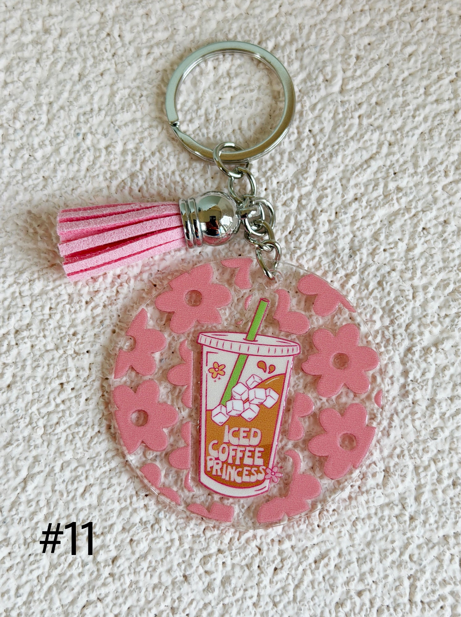 A pink acrylic keychain featuring a floral pattern, a pink tassel, and a clear round charm with a coffee cup illustration that reads 'ICED COFFEE PRINCESS'.