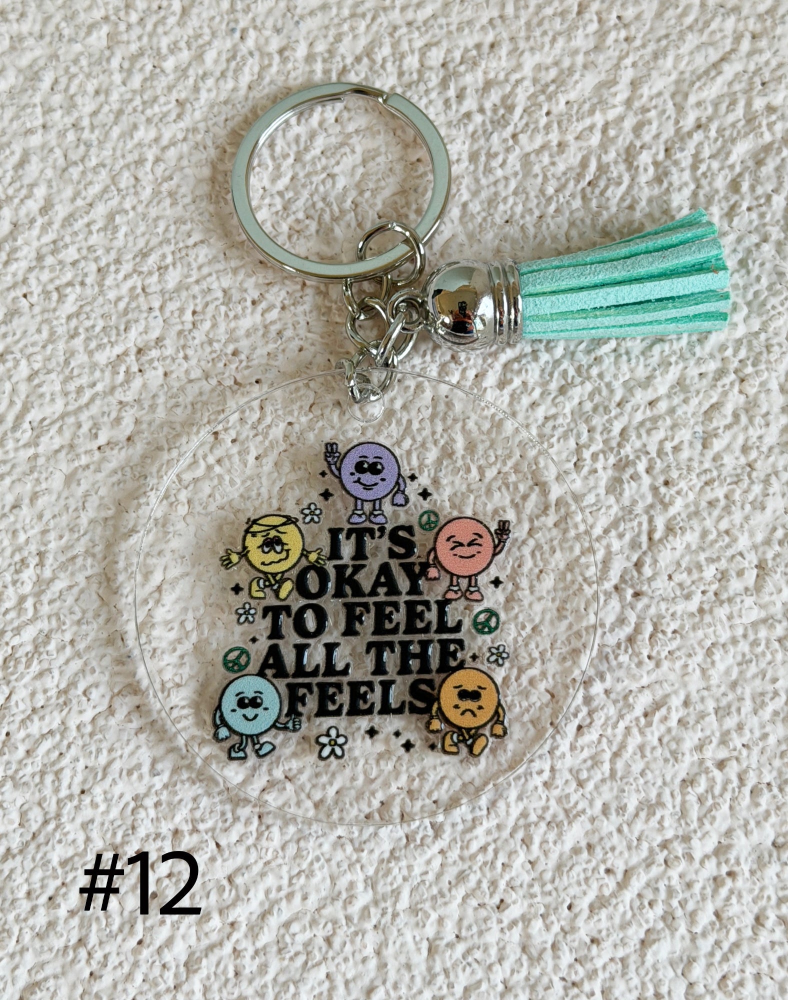 A clear acrylic keychain featuring a printed graphic of colorful cartoon characters and the text 'IT'S OKAY TO FEEL ALL THE FEELINGS' in black, attached to a mint green tassel and silver metal keyring, displayed on a textured white fabric background.