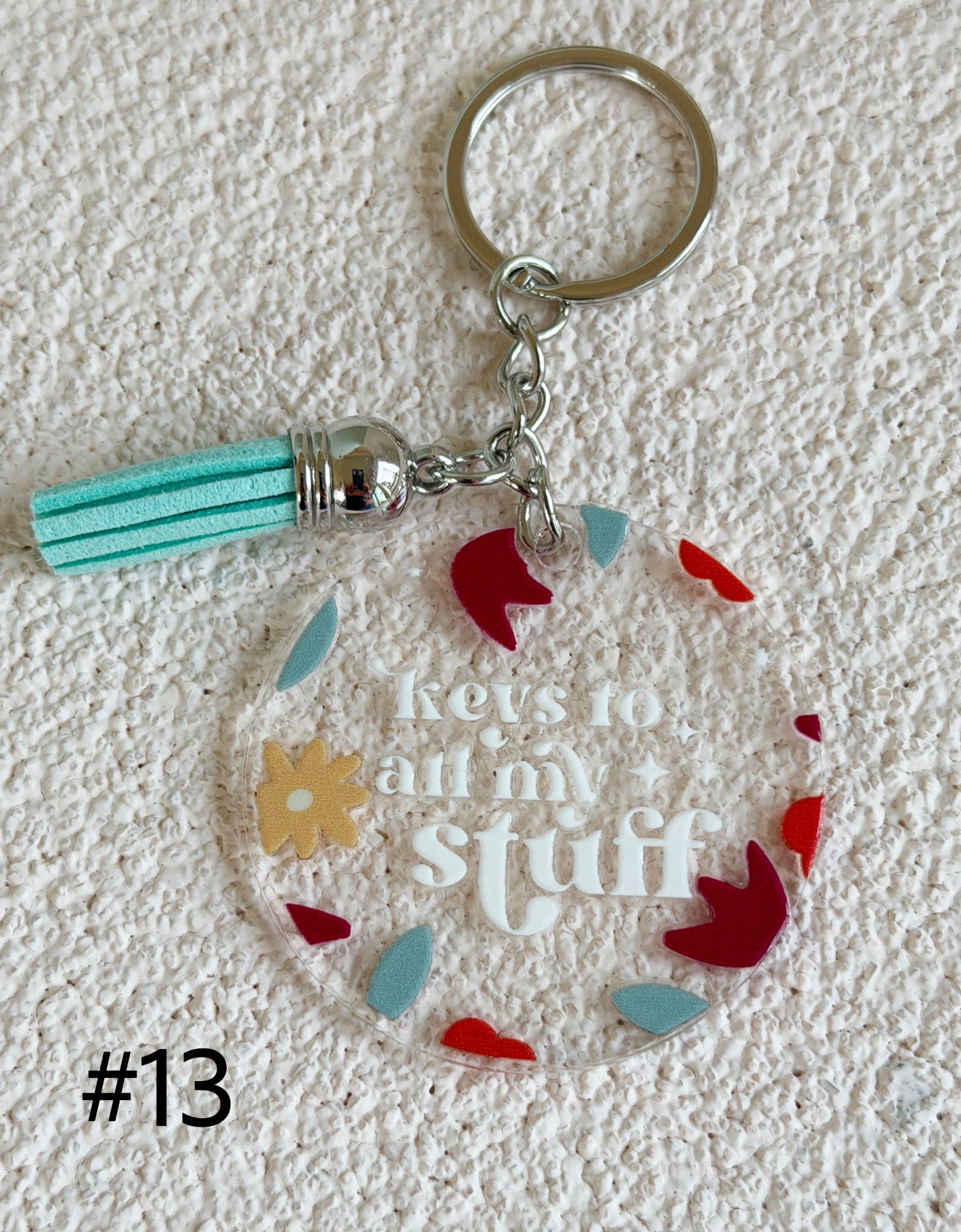 A clear acrylic keychain with a silver metal split ring and tassel, featuring the phrase 'keys to all my stuff' in white script, surrounded by small colorful decorative pieces in red, light blue, and yellow, displayed on a textured white background.