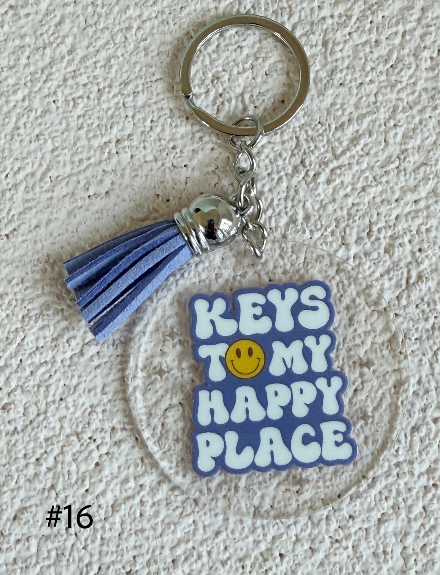 A square acrylic keychain charm with the text 'KEYS TO MY HAPPY PLACE' in white letters with a yellow smiley face in the center, attached to a lavender-blue tassel and a silver metal key ring, displayed on a textured white background.