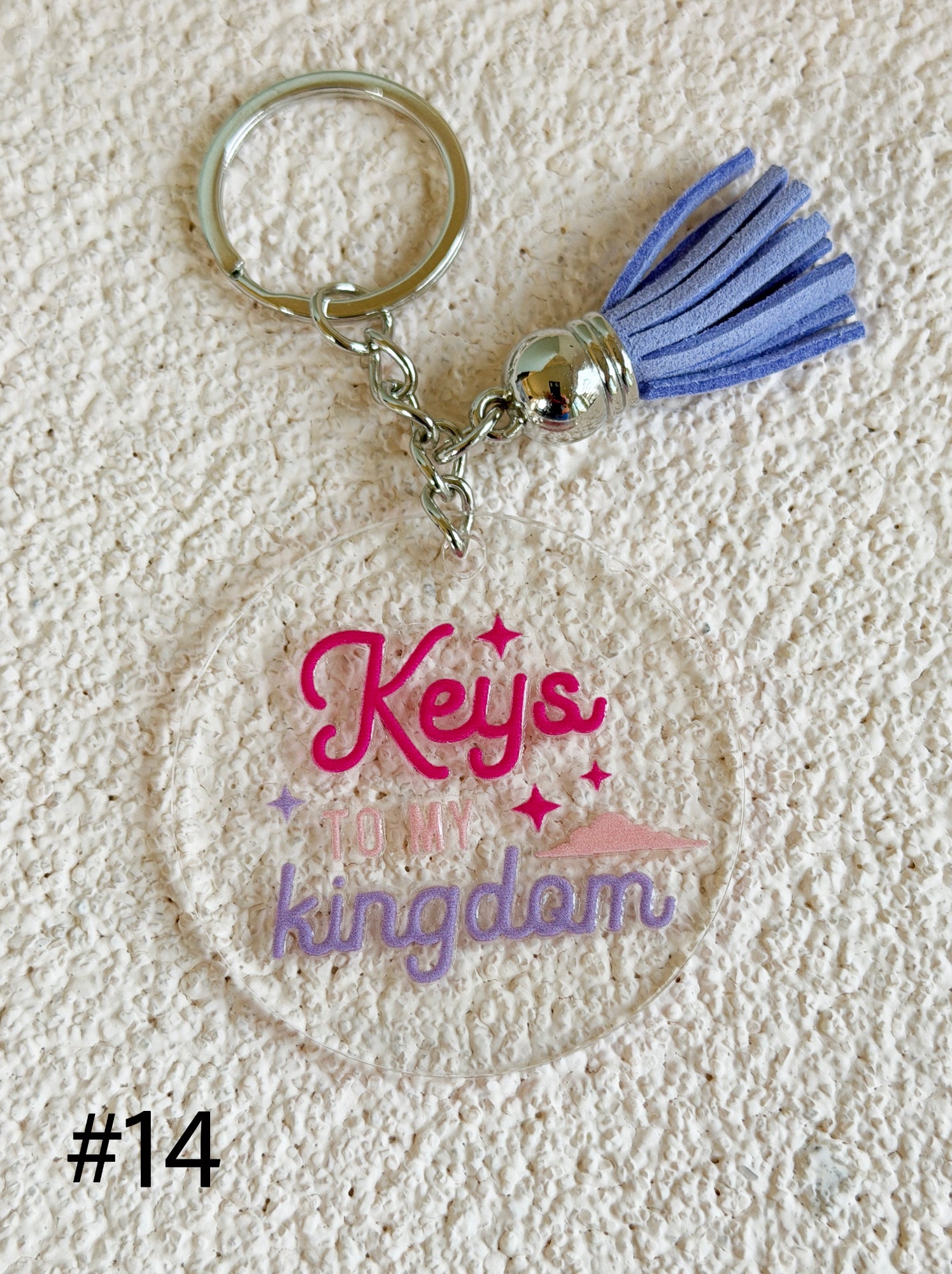 A round clear acrylic keychain featuring the text 'Keys to my kingdom' in pink and purple lettering, complete with a blue tassel and silver metal key ring, displayed on a textured beige background.