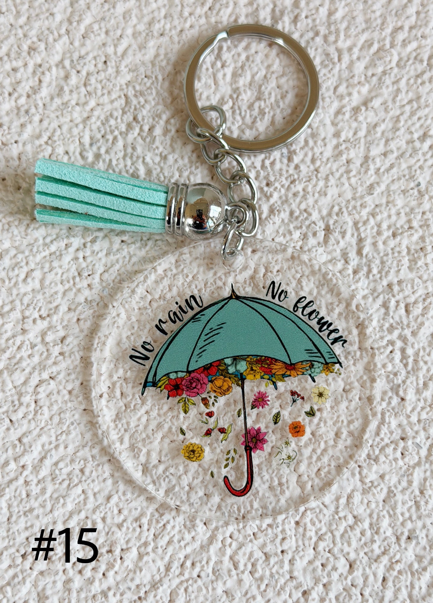 A round acrylic keychain charm featuring a mint green umbrella with colorful flowers and the words 'No rain, No flower' on a textured white background. The keychain includes a mint green tassel, silver metal rings, and a silver bead.