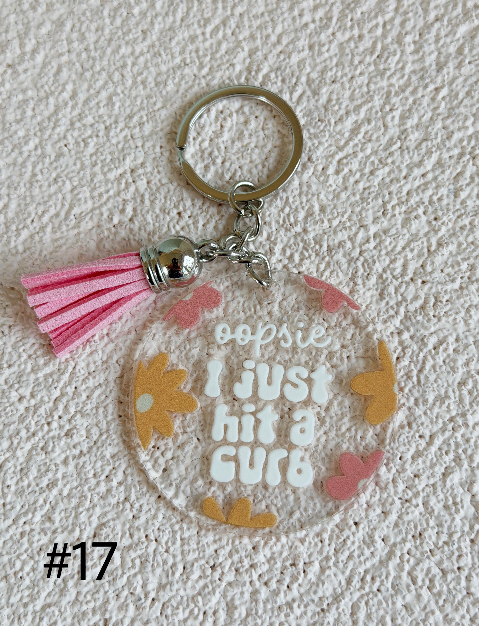 A clear acrylic keychain charm with a pink tassel and silver metal key ring, featuring the text 'oopsie I just hit a curb' in white lettering, surrounded by small yellow and pink flower and butterfly illustrations. The keychain is displayed on a textured white background with the text '#17' in the bottom-left corner.