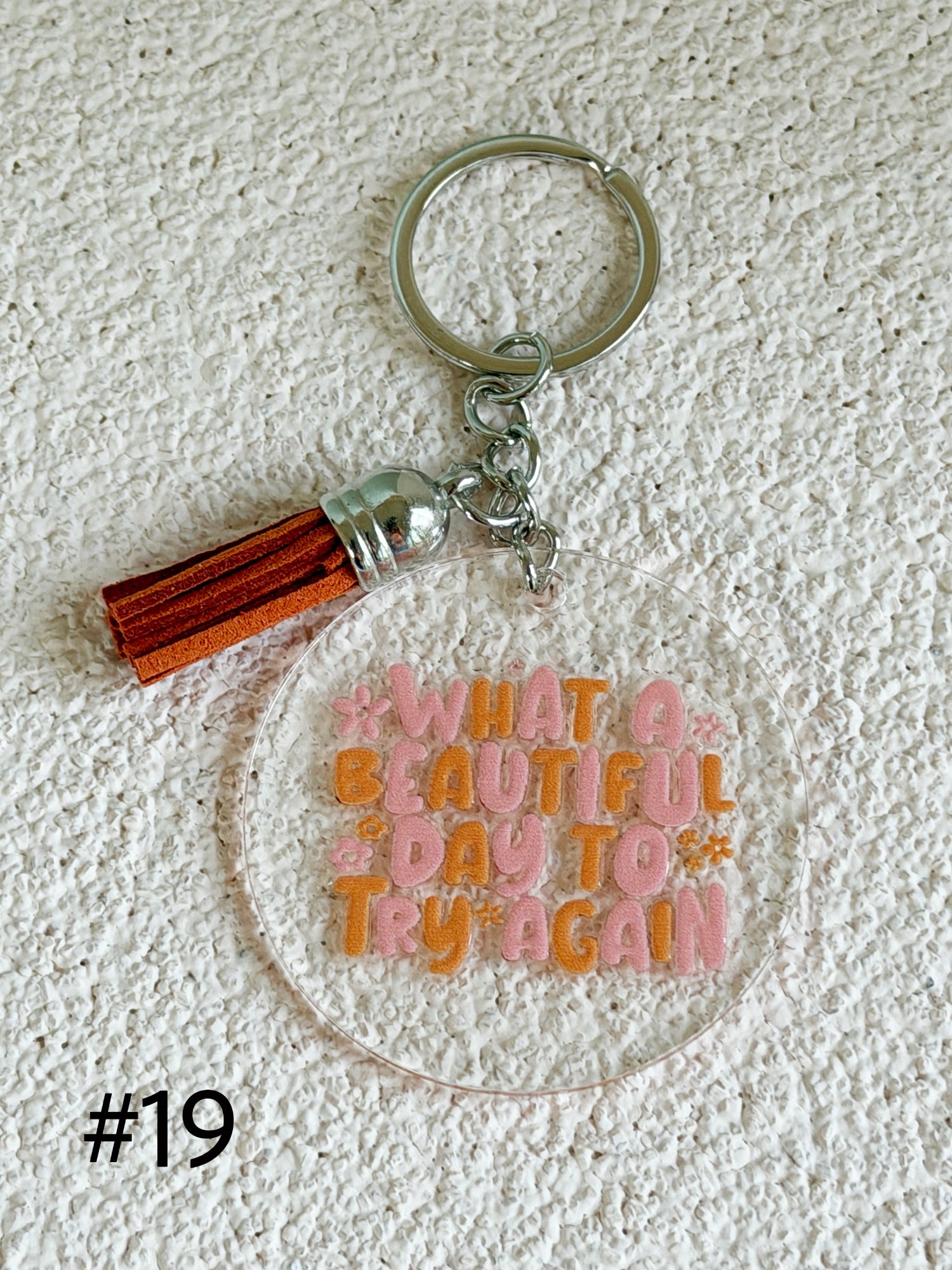 A clear acrylic keychain with a silver metal split ring and tassel charm, featuring the phrase 'WHAT A BEAUTIFUL DAY TO TRY AGAIN' in pink and orange lettering, displayed on a textured white background.
