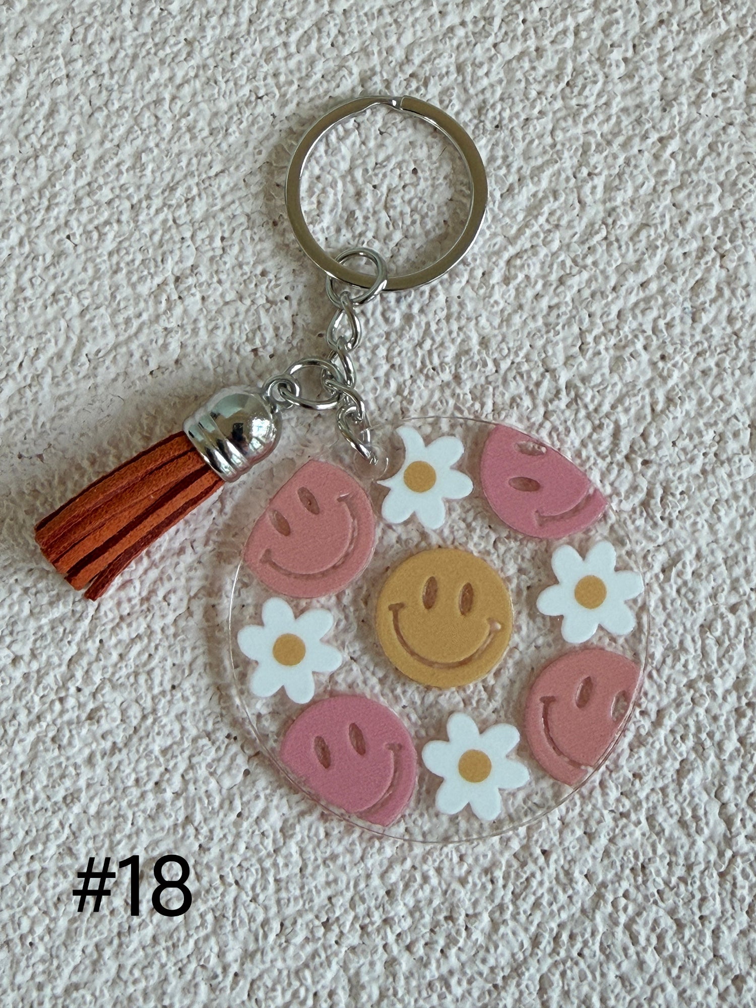A round clear acrylic keychain charm featuring pink smiley faces, white daisies, a yellow smiley face, and a brown leather tassel, displayed on a textured white background with '#18' text overlay.